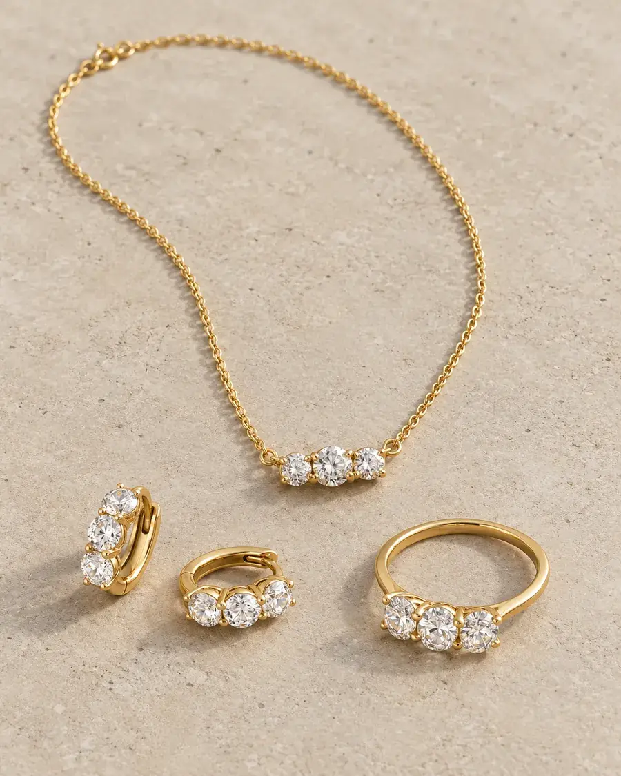 Gold three-stone necklace, huggie earrings and ring with round-cut stones on a stone surface